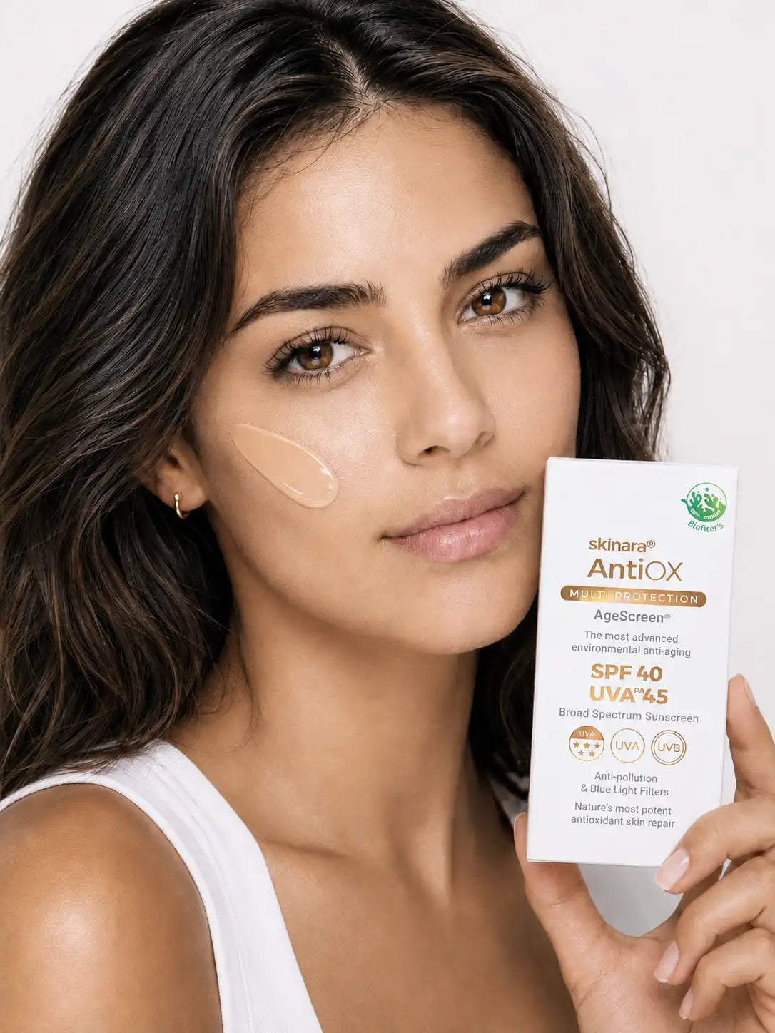 A woman with dark hair and olive skin shows a sunscreen smudge on her cheek and holds a box of skinara AntioX AgeScreen Broad Spectrum SPF 40 UVA/UVB sunscreen with high UVA anti-wrinkle. protection