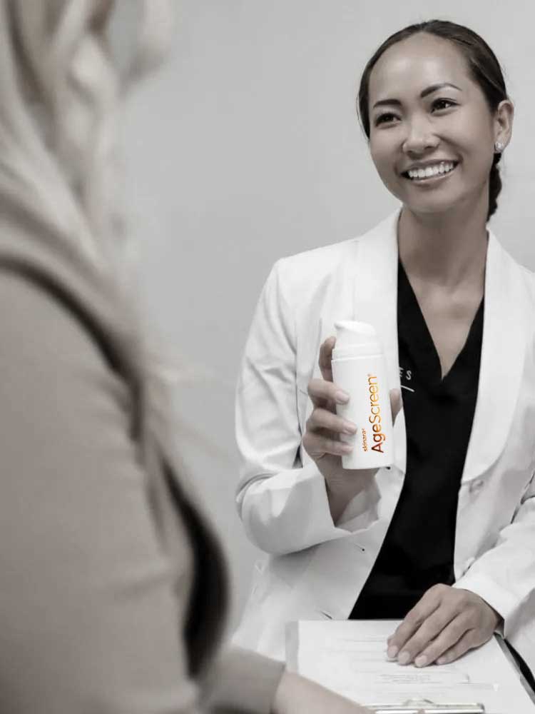 Woman dermatologist in a white coat holding a bottle of Skinara AgeScreen with a blurred background