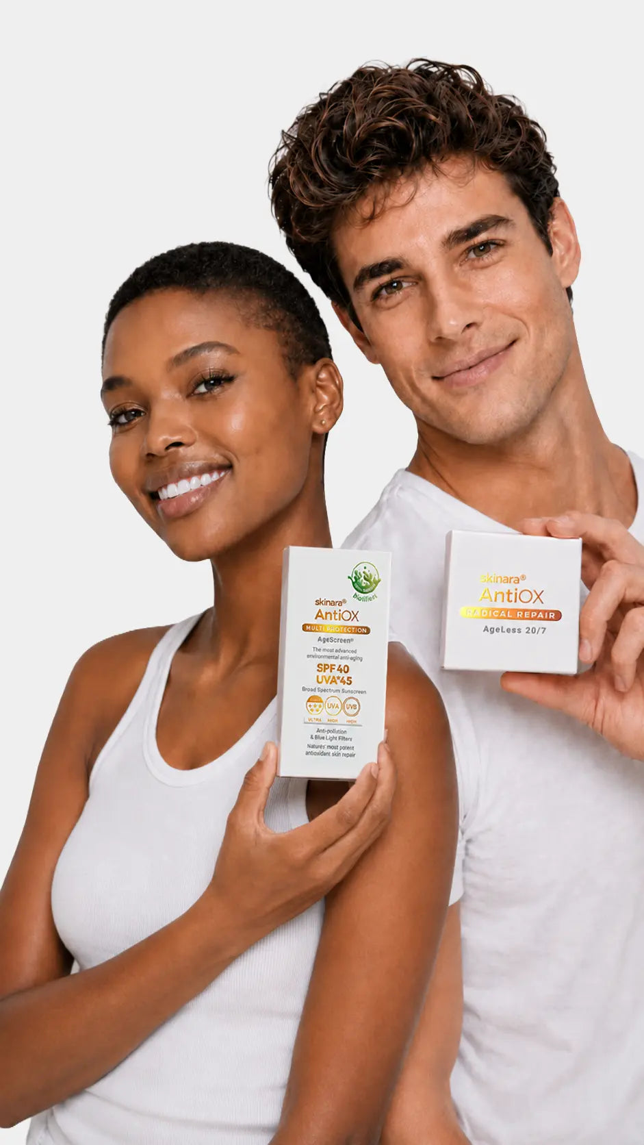 A smiling man and woman standing against a light gray studio background, each holding a small white box of skinara AntiOx Sunscreen.