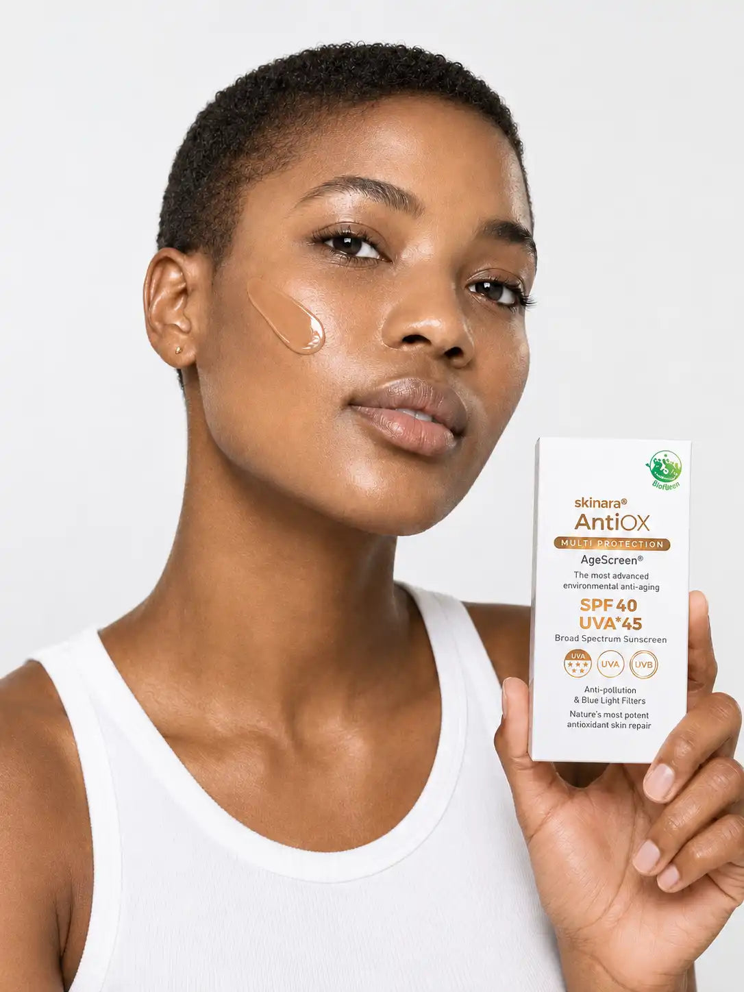 A woman with dark skin tone holds a box of Skinara Antiox AgeScreen SPF 40 UVA+45 sunscreen while applying a smear of the espresso shade to her cheek